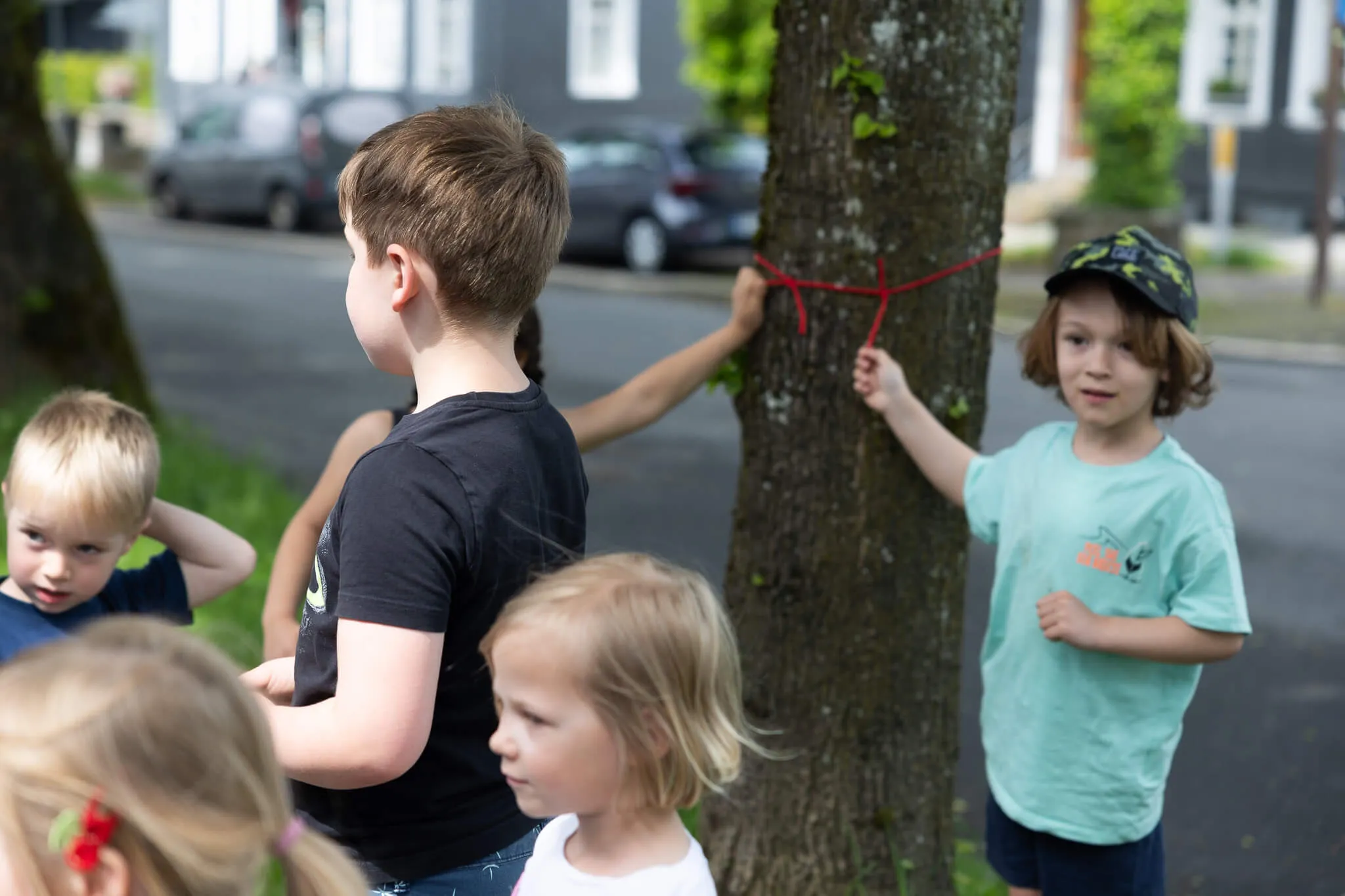 Kinder stehen im Kreis um einen Baum, ein Kind bindet ein rotes Band darum. Im Hintergrund sind Autos und Häuser zu sehen.