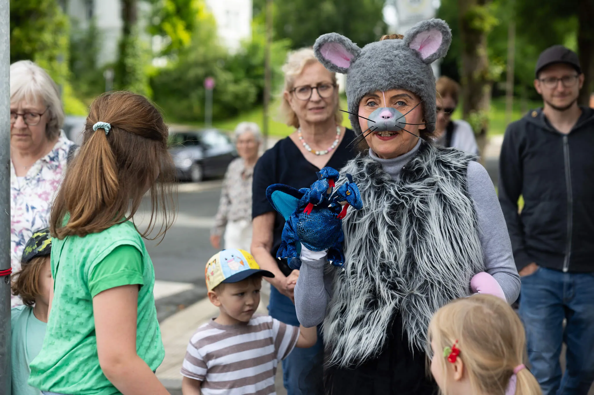 Eine Frau im Mäusekostüm spielt mit einem Handpuppen-Drachen vor Kindern und erwachsenen Zuschauern im Freien.