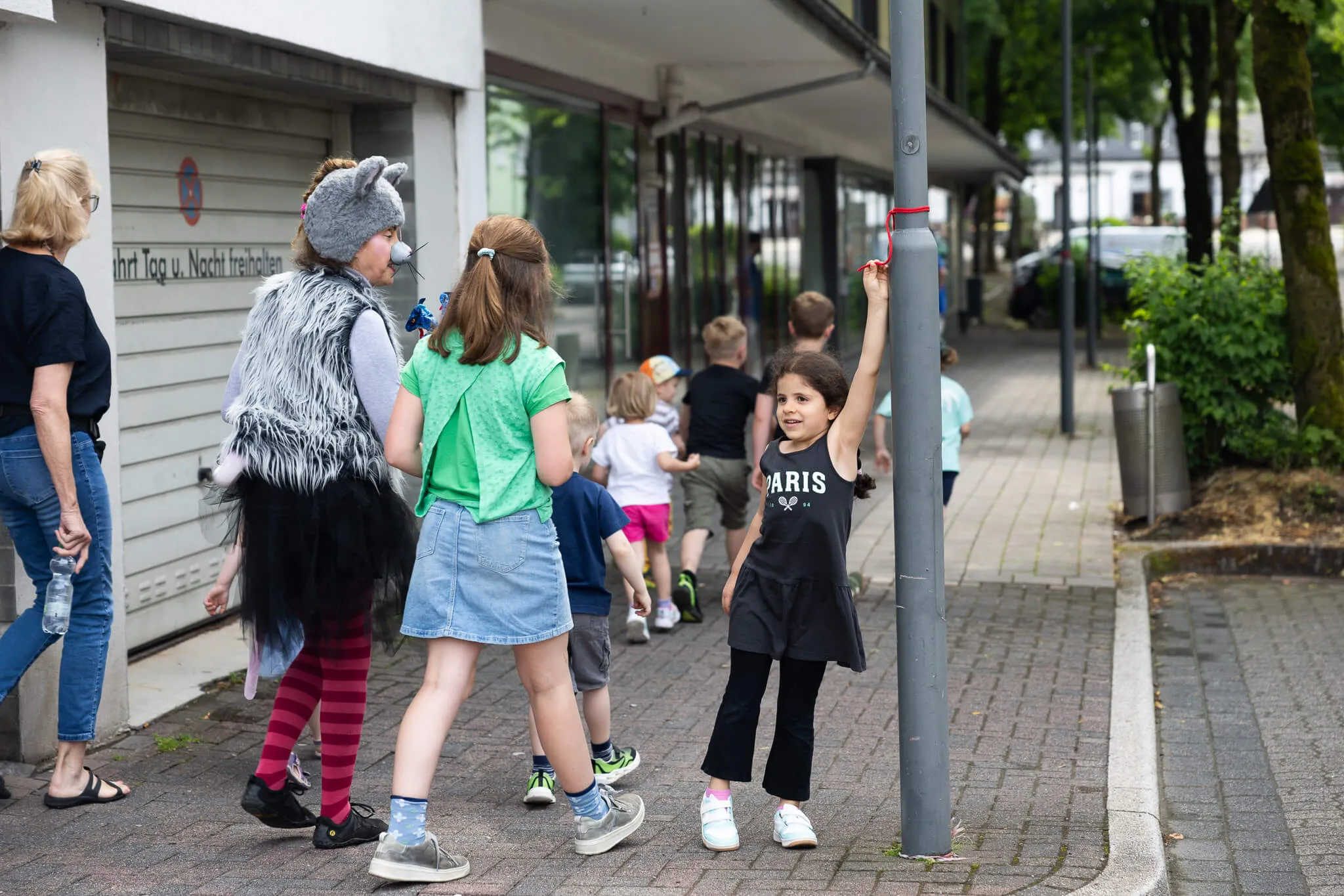 Kinder in Kostümen und Alltagskleidung gehen auf dem Gehweg neben einem Gebäude entlang, eines der Kinder hebt den Arm zu einem roten Band an einem Mast.