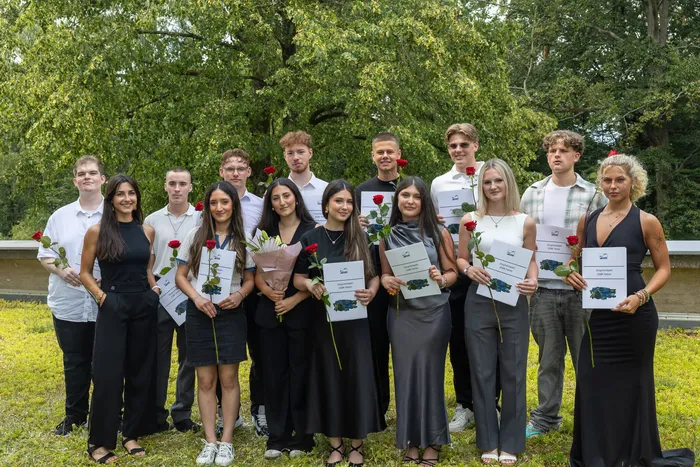 Gruppe von dreizehn jungen Erwachsenen, die an einer Abschlussfeier teilnehmen. Die meisten halten Urkunden und rote Rosen.
