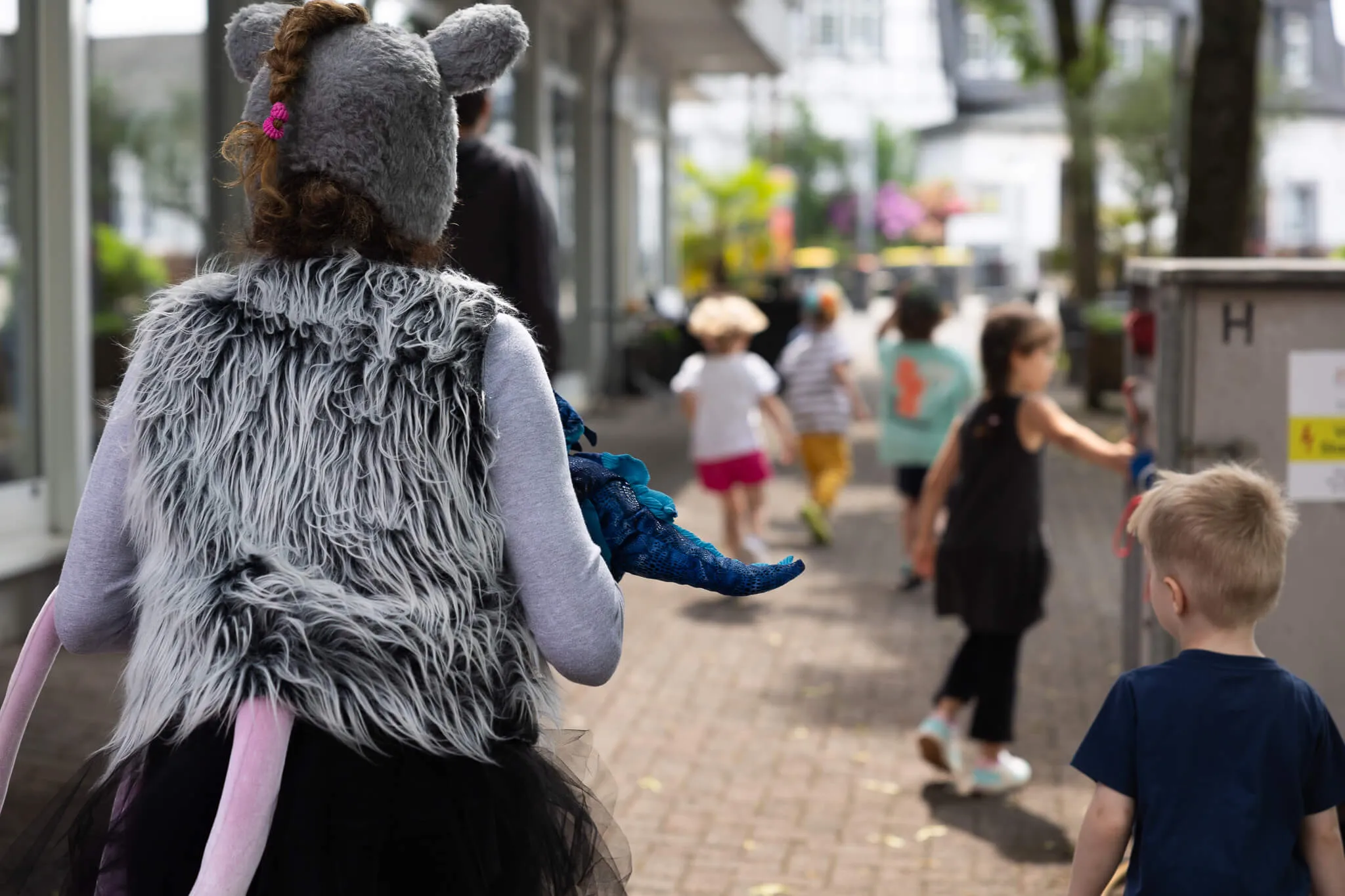 Eine Person in einem grau-weißem Tierkostüm mit langen Ohren führt eine Gruppe Kindergartenkinder durch eine Fußgängerzone. Sie hält ein blaues Stofftier in der Hand.