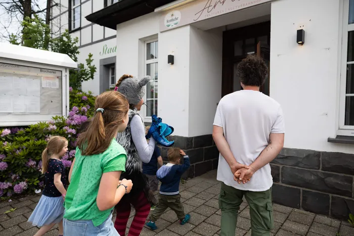 Eine Gruppe Kinder betritt mit einer erwachsenen Begleitperson ein Gebäude, vermutlich eine Stadtbücherei, während ein weiteres Kind mit einem blauen Stofftier in der Hand hinterherläuft.