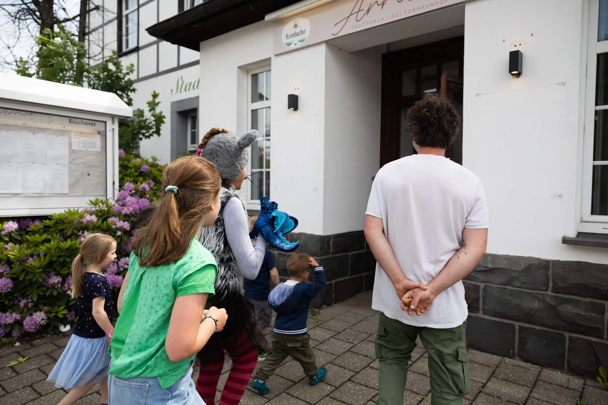 Eine Gruppe Kinder betritt mit einer erwachsenen Begleitperson ein Gebäude, vermutlich eine Stadtbücherei, während ein weiteres Kind mit einem blauen Stofftier in der Hand hinterherläuft.