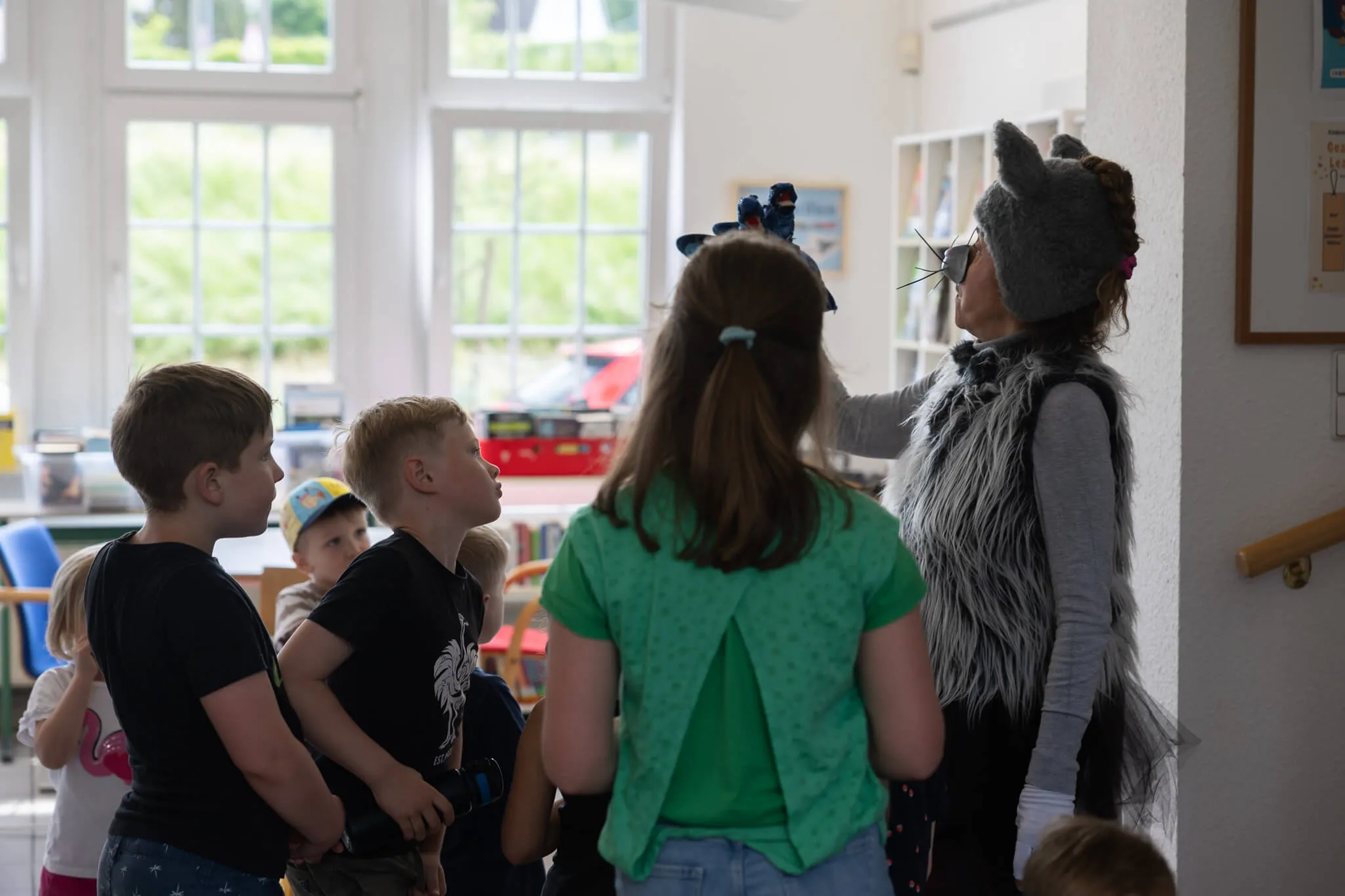 Eine Person mit Wolfskostüm interagiert mit einer Gruppe Kindergartenkinder in einem hellen Raum, vermutlich in einer Bücherei.