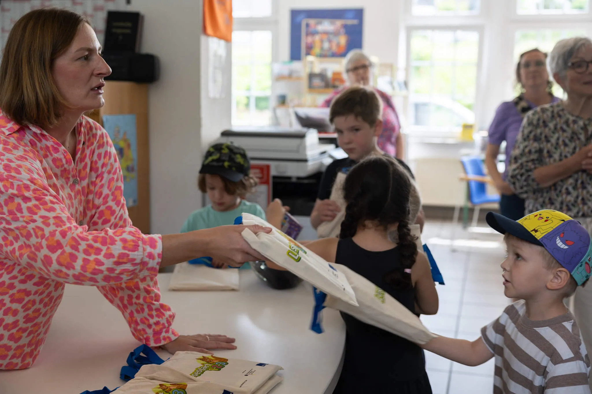 Eine Frau in einem gemusterten Kleid übergibt ein Kind an ein kleines Mädchen ein Geschenkpapier mit dem Logo der Stadtbücherei. Im Hintergrund sitzen und stehen weitere Kinder und zwei ältere Frauen.