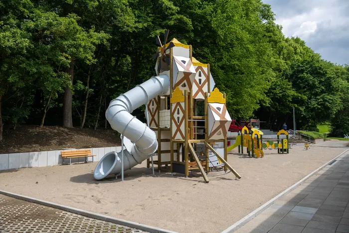 Ein Spielturm mit einer gewundenen Rutsche steht auf einem Sandplatz neben einem Gehweg und Bäumen.