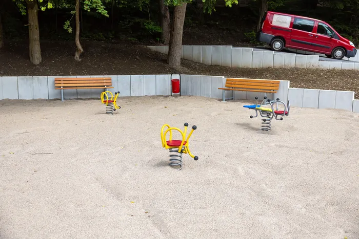 Sandkasten mit mehreren Federwippen und zwei Bänken, im Hintergrund ein roter Transporter.