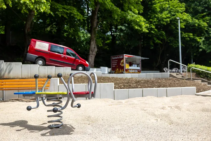 Ein Spielplatz mit einer blauen und roten Wippe im Vordergrund, im Hintergrund ein roter Lieferwagen und ein Imbissstand.