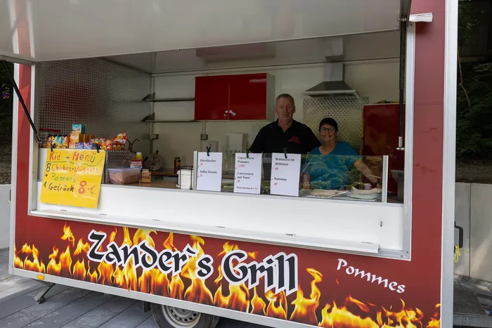 Ein Imbisswagen mit der Aufschrift „Zander’s Grill“ steht geöffnet, ein Mann und eine Frau stehen hinter der Theke. Im Angebot sind unter anderem Pommes und Chicken Nuggets.