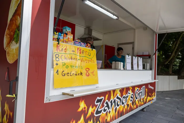 Gelbes Schild bewirbt ein Kindermenü mit 8 Chicken-Nuggets, Pommes und einem Getränk für 8 Euro vor einem Imbisswagen mit der Aufschrift „Zander’s Grill“.