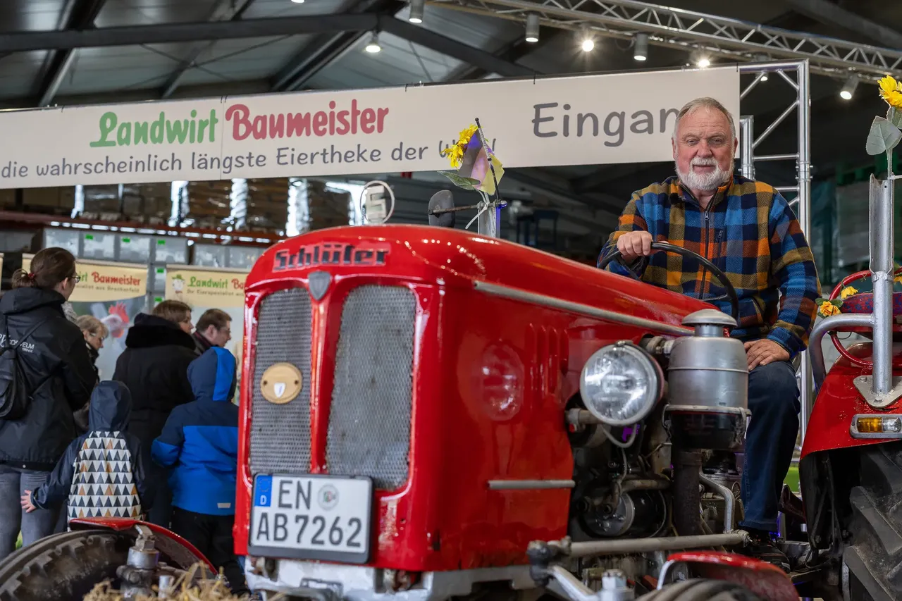 Ein älterer Mann mit Bart sitzt lächelnd auf einem roten Oldtimer-Traktor vor einer belebten Markthalle. Im Hintergrund ein Banner mit der Aufschrift „Landwirt Baumeister“ und „Eingang“.