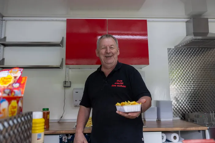 Ein Mann lächelt in einem Imbisswagen und hält eine Portion Pommes Frites auf einem weißen Tablett bereit.
