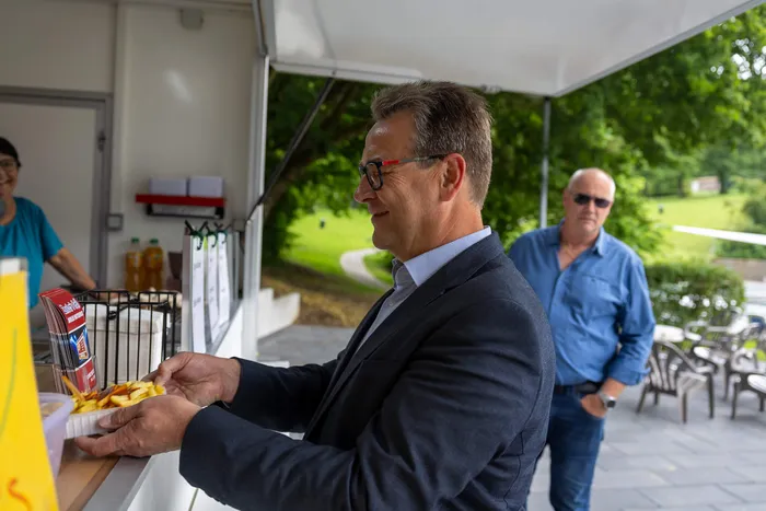 Ein Mann in Anzug und Brille nimmt Pommes Frites aus einer Papiertüte entgegen, während im Hintergrund ein anderer Mann mit Sonnenbrille steht. Im Hintergrund ist eine grüne Landschaft zu sehen.
