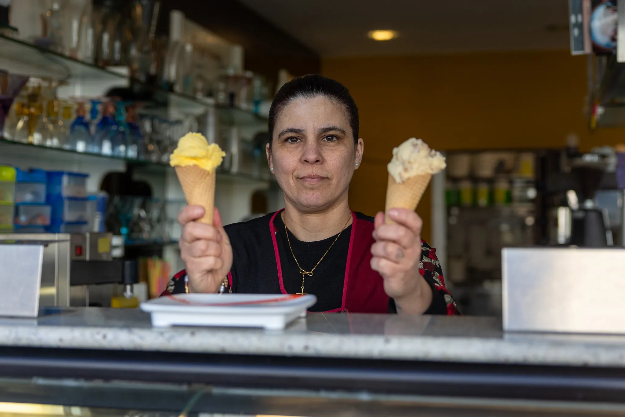Mitarbeiterin präsentiert zwei Waffeleis vor der Theke eines Eiscafés mit Regalen voller Gläser und Behälter im Hintergrund.