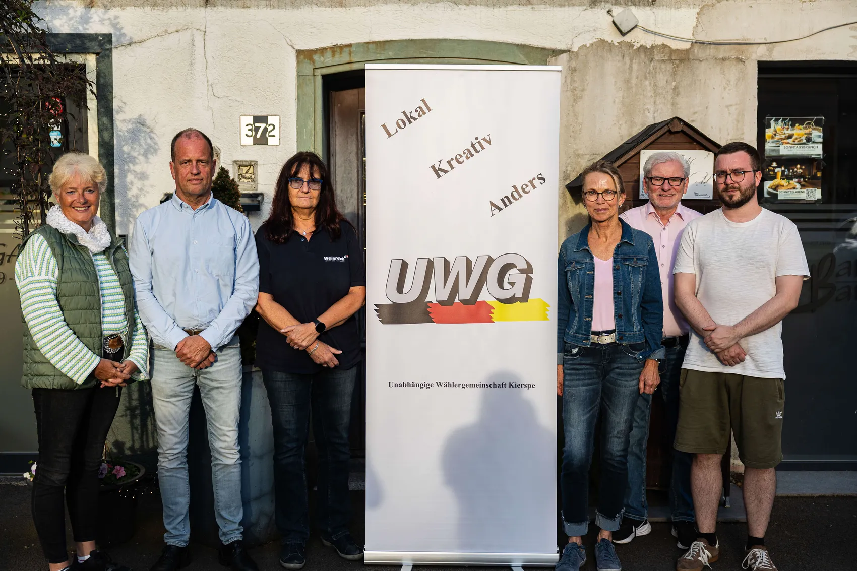 Sechs Personen stehen vor einem Gebäude mit einem Banner der UWG Kierspe. Das Banner zeigt die Farben der deutschen Flagge und den Schriftzug „Lokal, Kreativ, Anders“.