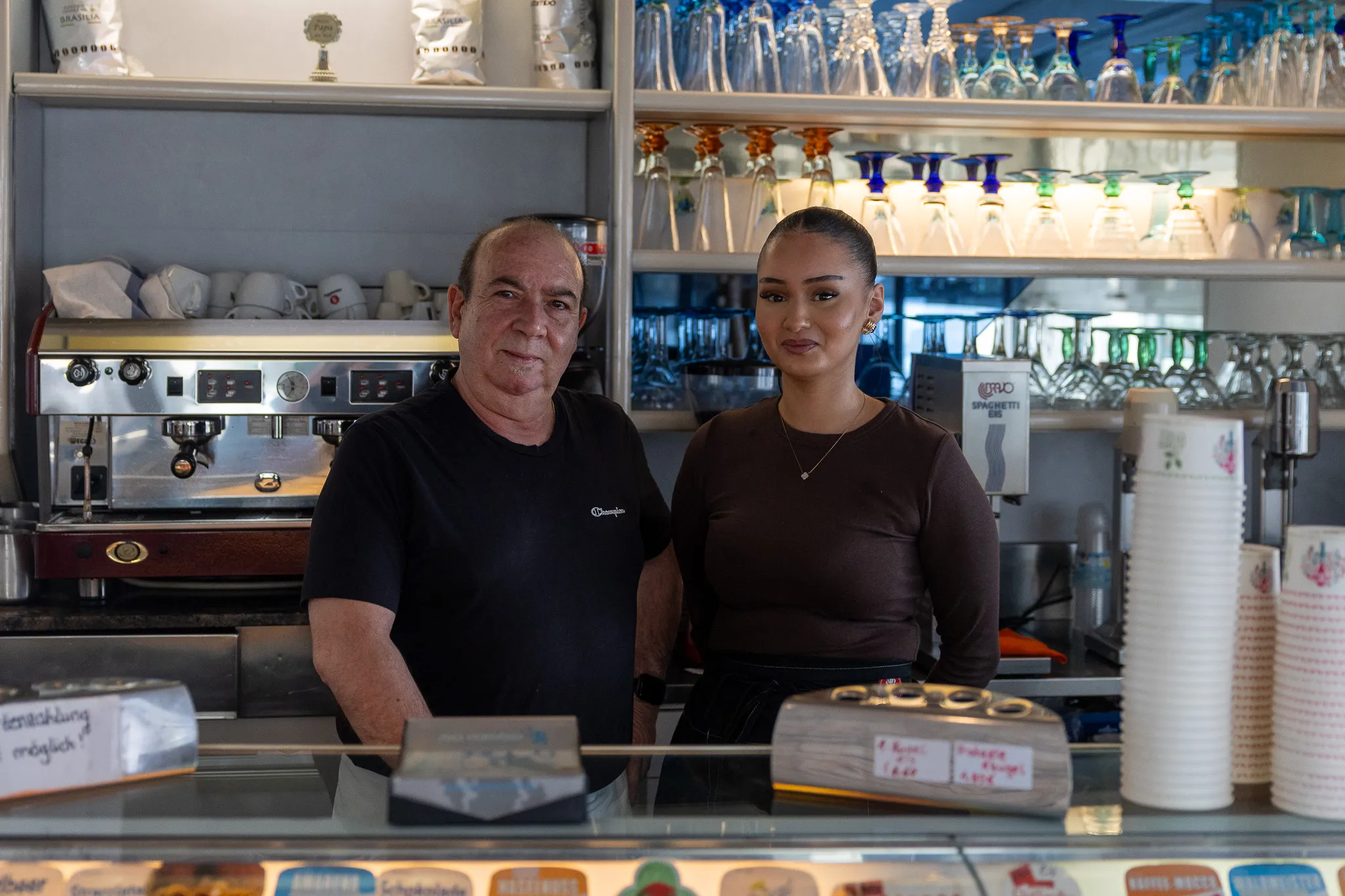 Ein Mann und eine junge Frau stehen hinter einer Eistheke in einem Café. Im Hintergrund sind Regale mit Gläsern und Kaffeemaschinen zu sehen.
