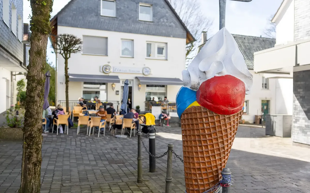 Ein großes, bemaltes Eis auf Waffel steht im Vordergrund vor dem Eiscafé Venezia in Breckerfeld, wo Gäste an Tischen sitzen.