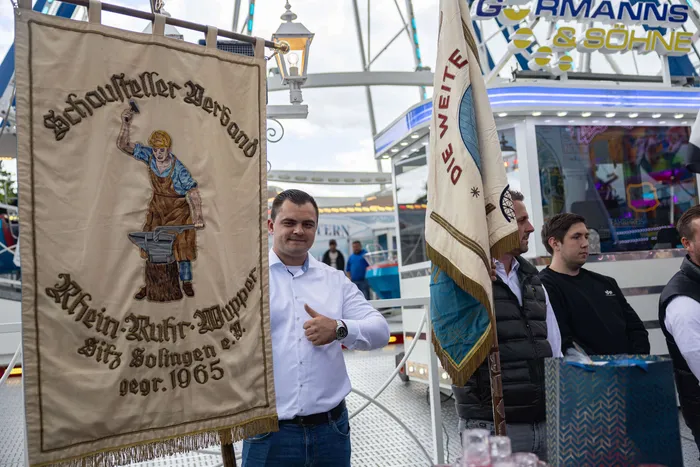Bürgermeister Armin Kibbert hält eine historische Vereinsfahne mit einem Wappen und einer Darstellung eines Schmieds, während er den Daumen nach oben zeigt. Im Hintergrund sind weitere Fahnen und ein belebter Kirmesplatz zu sehen.