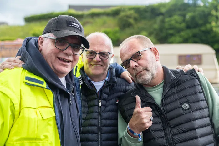 Drei Männer posieren eng zusammen für ein Foto, der Mann rechts zeigt Daumen hoch. Im Hintergrund ist ein Wohnmobil zu sehen.