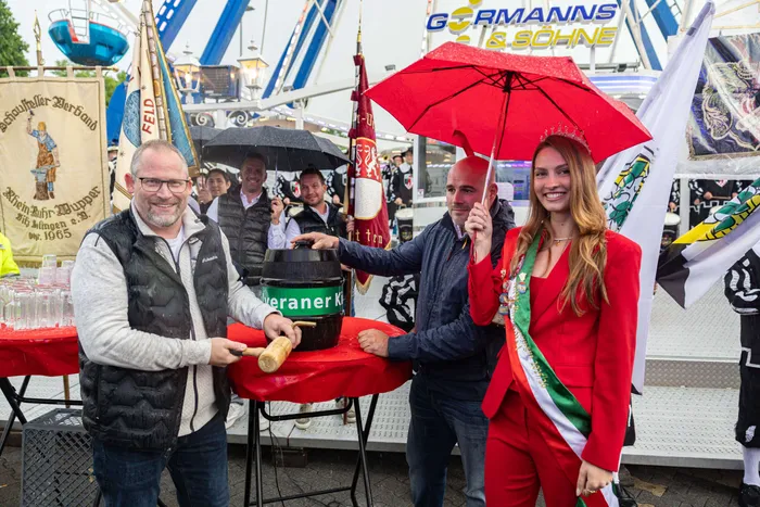 Bürgermeister Armin Kibbert zapft bei der Eröffnung der Halveraner Kirmes ein Bierfass an, während eine junge Frau mit einem roten Band und einem Regenschirm daneben steht. Im Hintergrund sind Fahrgeschäfte und Buden zu sehen.