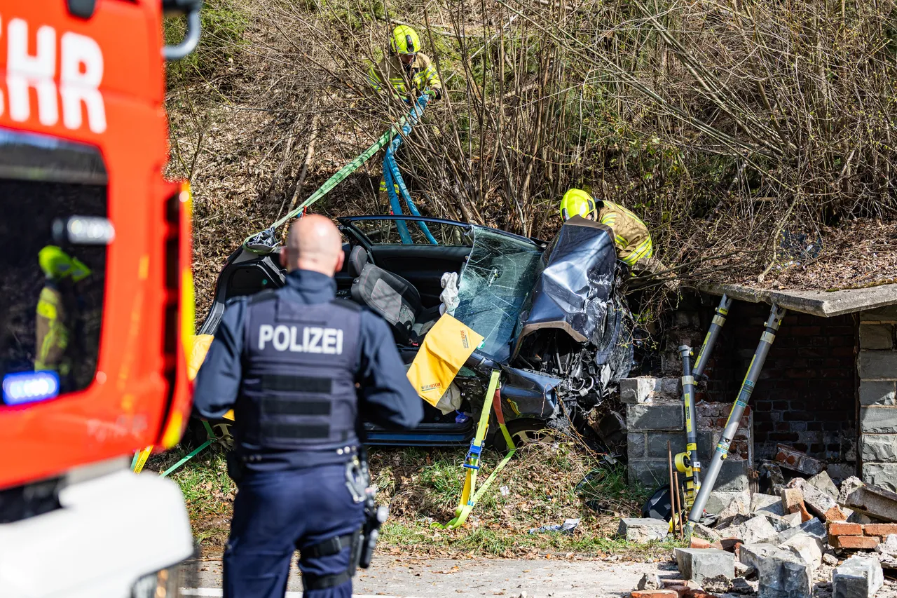 Ein stark beschädigtes Auto ist gegen einen Baum geprallt, Einsatzkräfte der Feuerwehr und Polizei sind vor Ort. Trümmerteile liegen auf dem Boden.
