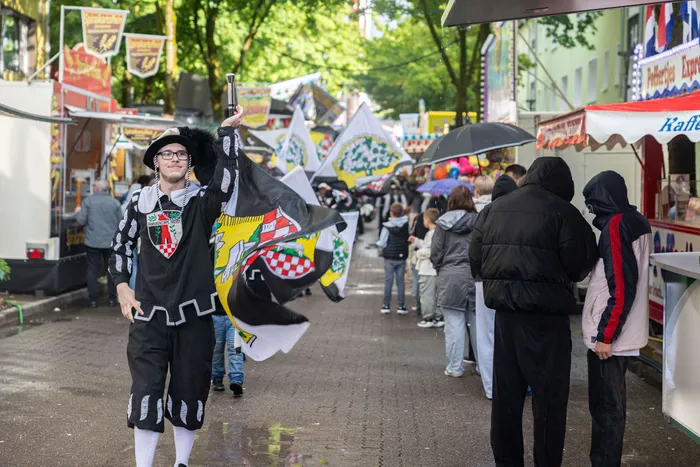 Ein Mann in historischer Tracht mit Wappen auf der Kleidung hebt ein Glas bei der Eröffnung der Kirmes. Im Hintergrund sind Buden und Menschen zu sehen.