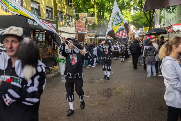 Bürgermeister Armin Kibbert trägt eine traditionelle Tracht und hält eine Fahne während der Eröffnung der Halveraner Kirmes. Im Hintergrund sind weitere Kirmesbesucher und Essensstände zu sehen.