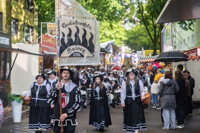 Eine Gruppe von Menschen in traditioneller Tracht zieht während der Eröffnung der Kirmes in Halver durch die Straßen. Ein Mann trägt eine Fahne mit der Aufschrift „Landsknechte Halver 1958“.