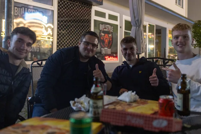 Vier junge Männer sitzen an einem Tisch im Freien, lächeln und zeigen Daumen hoch. Im Hintergrund ist ein Geschäft mit dem Schriftzug „Bullstyle Tattoo“ zu sehen.