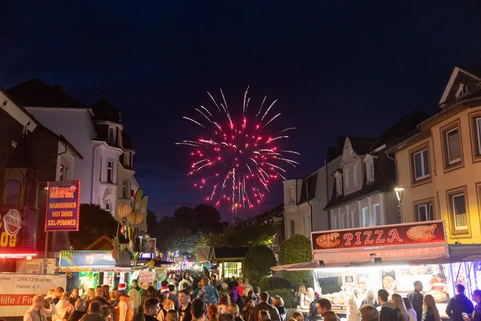 Rotes Feuerwerk über einer belebten Kirmes mit Essensständen und Menschenansammlungen zwischen Häusern.