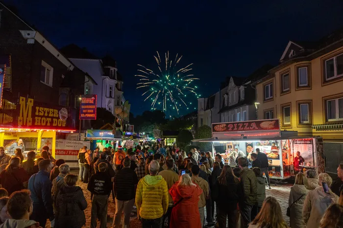 Feuerwerk in Form eines grünen Herzens erhellt den Nachthimmel über einer belebten Kirmes mit Imbissständen und Menschenansammlungen.