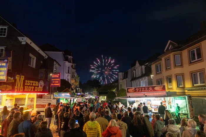 Menschenmenge auf einer Kirmes blickt zu einem Feuerwerk am dunklen Nachthimmel auf. Im Vordergrund Essensstände mit Beleuchtung.