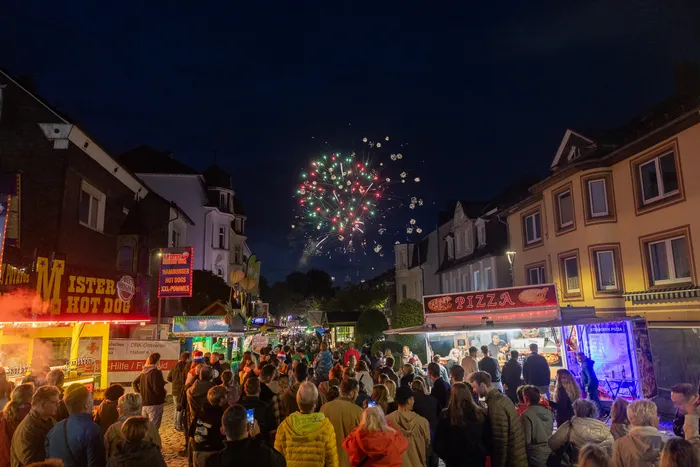 Feuerwerk in den Farben Rot, Grün und Weiß erhellt den dunklen Himmel über der belebten Halveraner Kirmes mit Essensständen und Menschenmengen.
