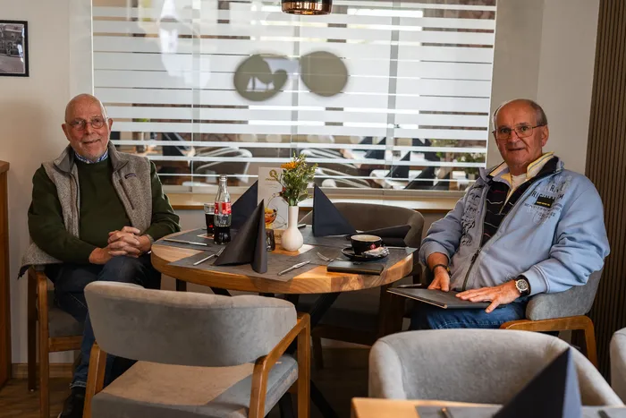Zwei ältere Männer sitzen an einem Tisch in einem hellen Restaurant und unterhalten sich. Auf dem Tisch stehen Getränke und eine kleine Blumengesteck.