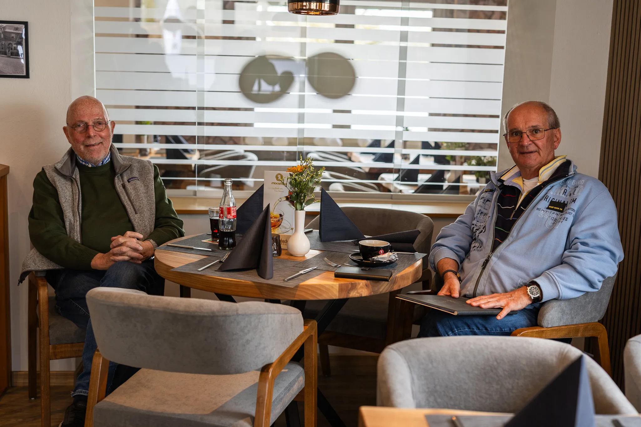 Zwei ältere Männer sitzen an einem Tisch in einem hellen Restaurant und unterhalten sich. Auf dem Tisch stehen Getränke und eine kleine Blumengesteck.