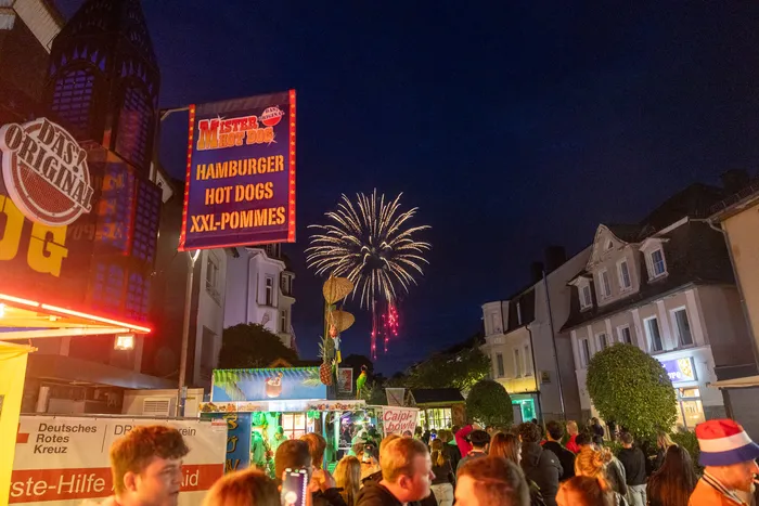 Feuerwerk erhellt den Nachthimmel über belebter Kirmes mit Imbissständen und Menschenansammlungen.