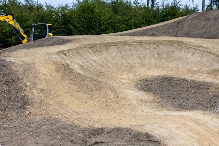 Eine geschwungene Erdbahn in einem neuen Bikepark, im Hintergrund ist ein gelber Bagger zu sehen.