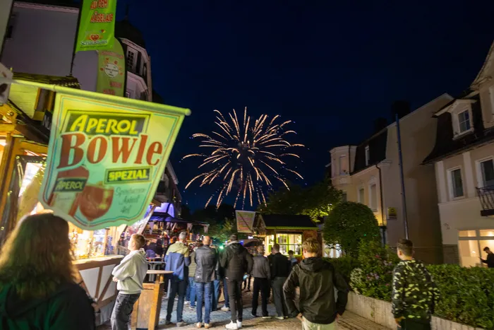 Feuerwerk erhellt den Nachthimmel über einer belebten Kirmes mit Besuchern vor einem Getränkestand mit der Aufschrift „Aperol Bowle Spezial“.
