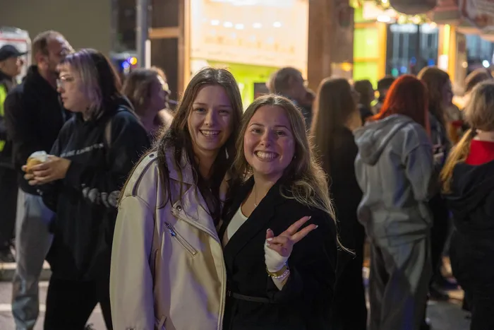 Zwei junge Frauen lächeln in die Kamera, im Hintergrund eine Menschenmenge und Lichter der Kirmes.