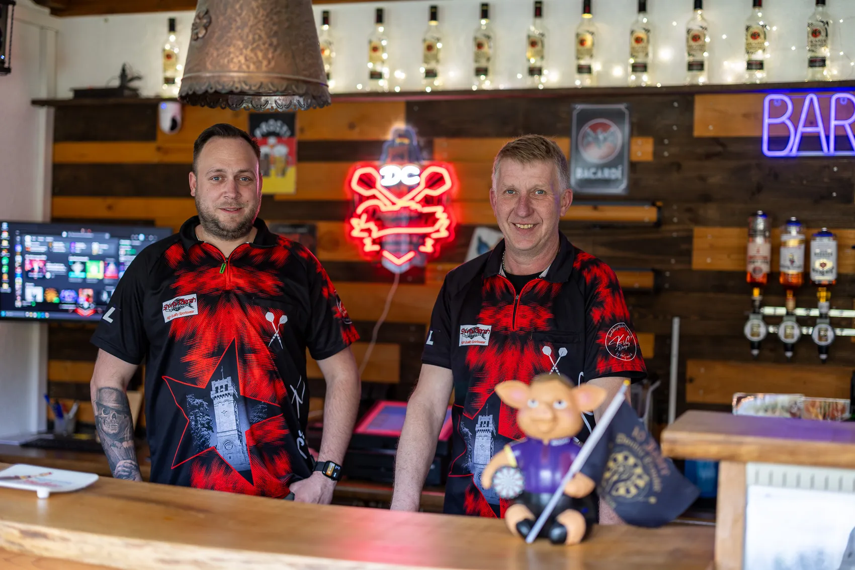 Zwei Männer in karierten Trikots stehen vor einer Bar mit Getränken im Hintergrund. Links Lars Kartschewski, rechts Martin Bartelt, Vorsitzende des Vereins.