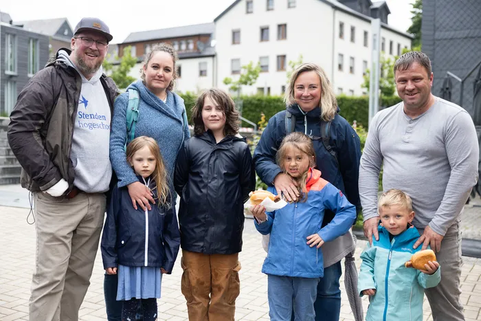 Eine Gruppe von sieben Personen, darunter zwei Kinder, steht lächelnd vor einer städtischen Kulisse. Einige halten Brötchen in den Händen.