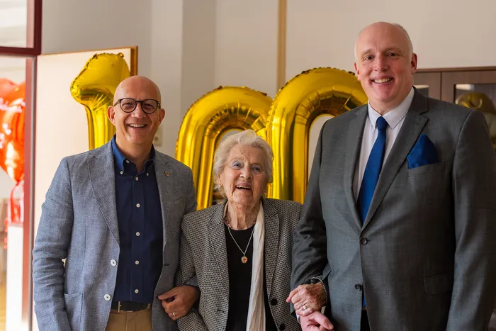 Emmi Seifert lacht gemeinsam mit Ralf Schwarzkopf und Christoph Weiland vor großen goldenen Luftballons, die die Zahl 100 bilden.