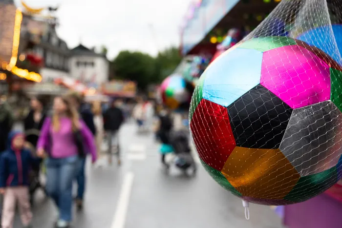 Bunter, geometrisch gemusterter Luftballon im Vordergrund vor verschwommenem Kirmestrubel mit Menschen und Fahrgeschäften.