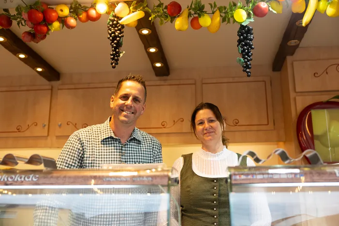 Mann und Frau lächeln in einer Konditorei oder Schokoladenladen. Über ihnen hängen dekorative Früchte und Weintrauben.