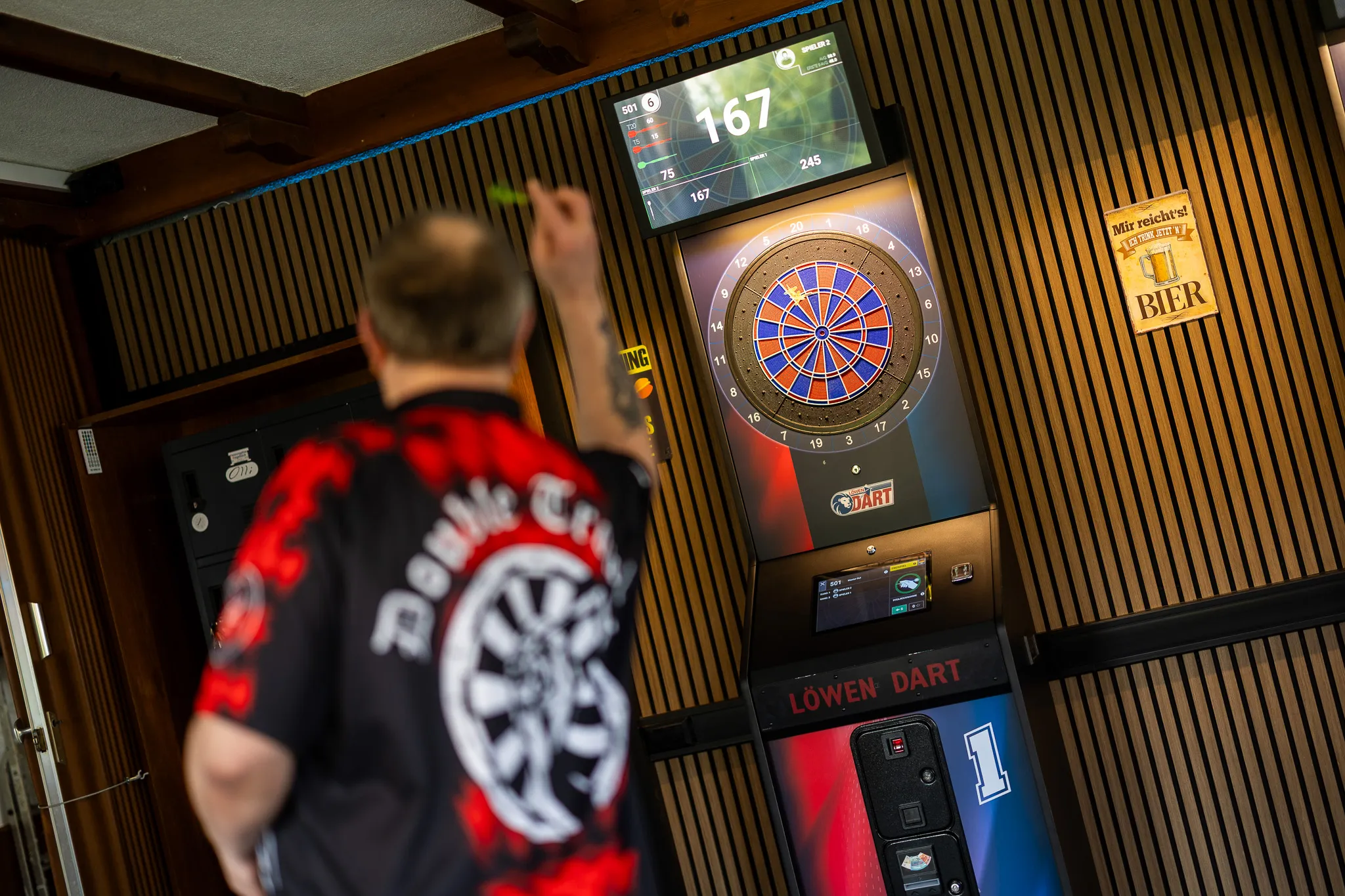 Ein Mann wirft einen Dart auf eine elektronische Dartscheibe in einem Vereinsheim. Im Hintergrund sind weitere Dartautomaten und ein Werbeplakat für Bier zu sehen.