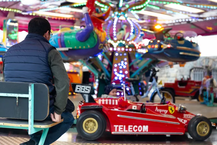 Ein Mann sitzt auf einer Bank vor einem bunten Karussell mit Fahrzeugen wie Rennwagen und Motorrädern. Im Hintergrund ist die beleuchtete Kirmes zu sehen.