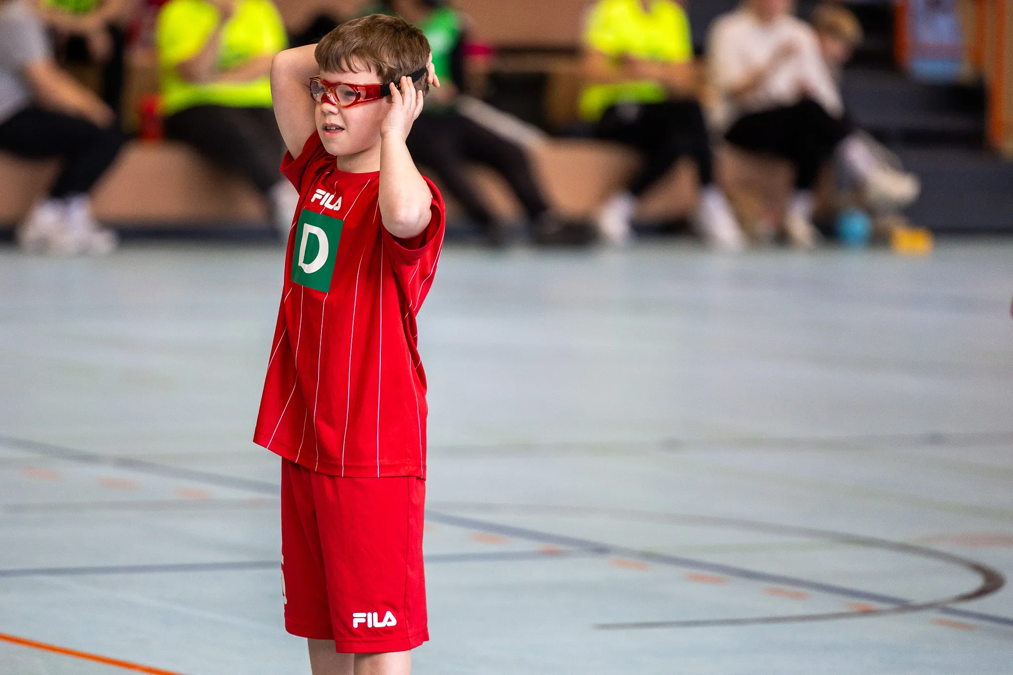 Ein Kind in roter Sportkleidung mit Schutzbrille steht auf einem Hallenboden und hält sich den Kopf. Im Hintergrund sind verschwommene Personen und Sportgeräte zu sehen.