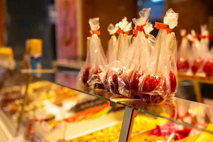 Reihe roter Zuckerstangen in durchsichtigen Plastiktüten auf einem Verkaufstresen vor einer unscharfen Kirmes-Kulisse.
