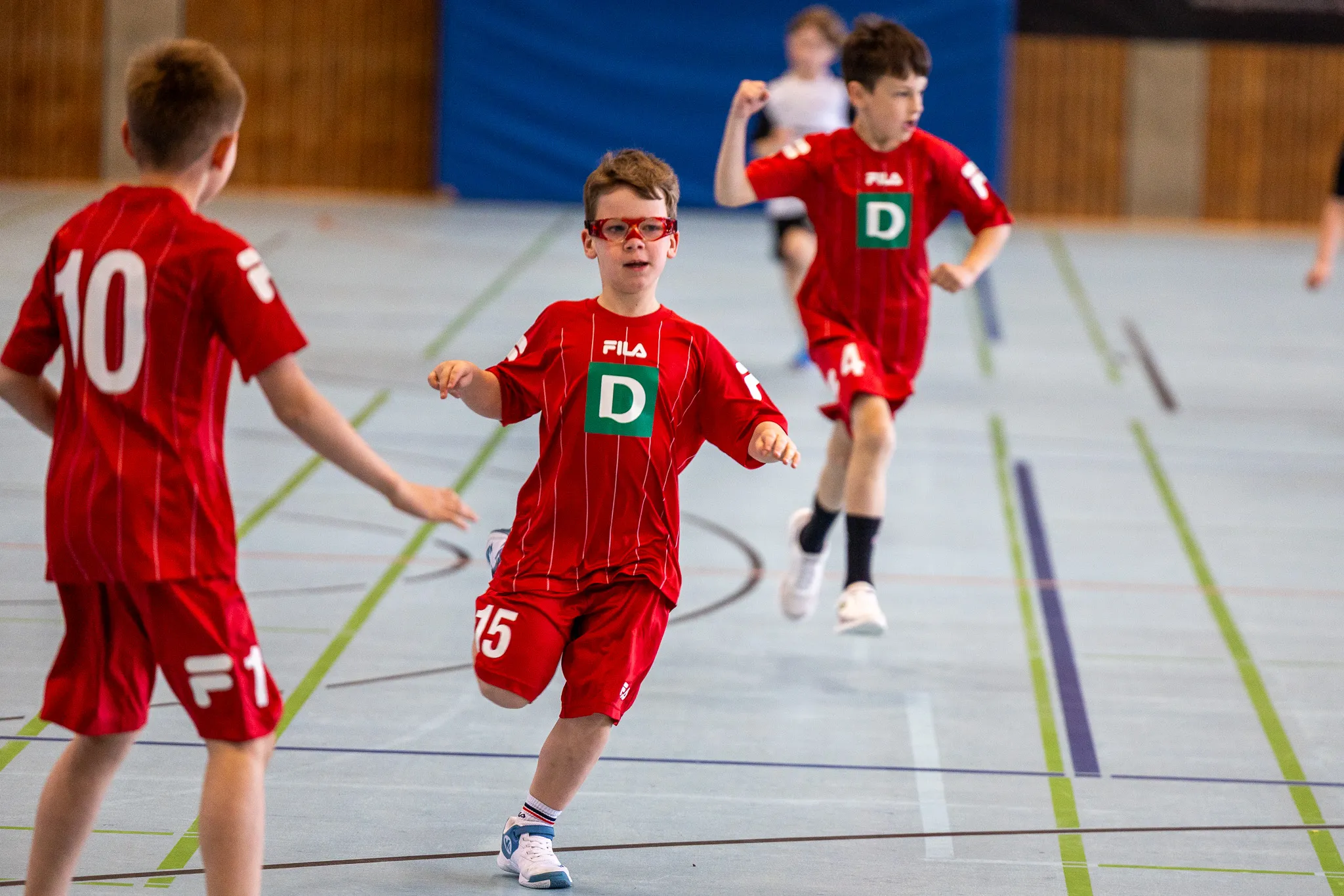 Junge mit Brille in rotem Handballtrikot rennt während eines Spiels in einer Sporthalle. Im Hintergrund weitere Spieler und Zuschauerränge.
