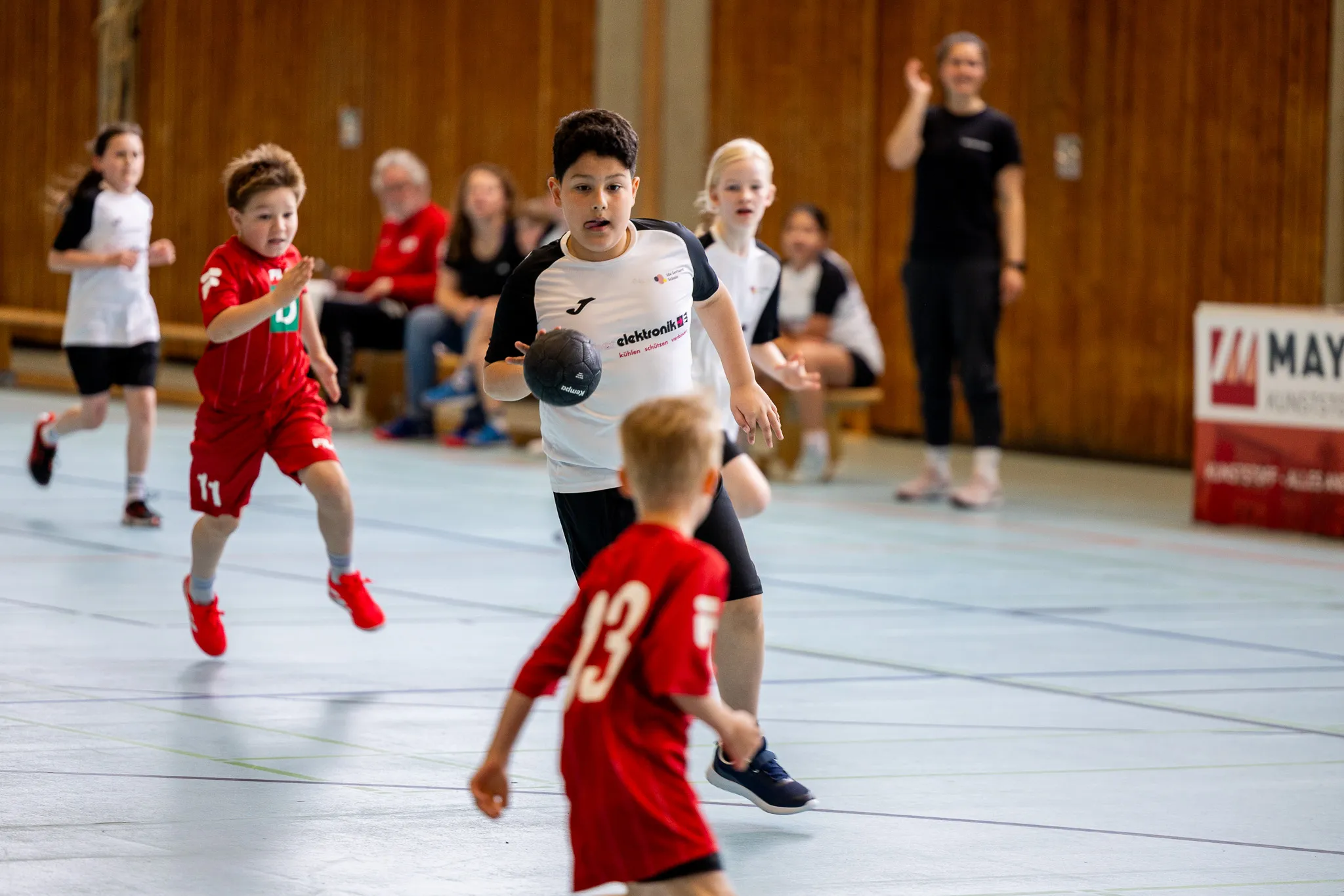 Jungen spielen Handball in einer Sporthalle. Ein Spieler mit dem Ball wird von einem Gegenspieler verfolgt, während andere Kinder im Hintergrund stehen.